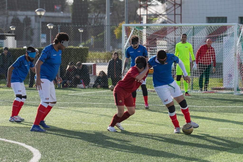 Campionato Europeo 2023 - 2024. Tappa Roma. In foto, un calciatore, che indossa un completo con pantaloncini cloro bianco e maglietta color azzurro, sta calciando il pallone. Accanto a lui c'è un calciatore che indossa un completo pantaloncini e maglietta color rosso, che tenta di spingerlo allontanandolo dal pallone. Dietro sono presenti anche due calciatori che indossano completo con pantaloncini color bianco e maglietta color azzurro.