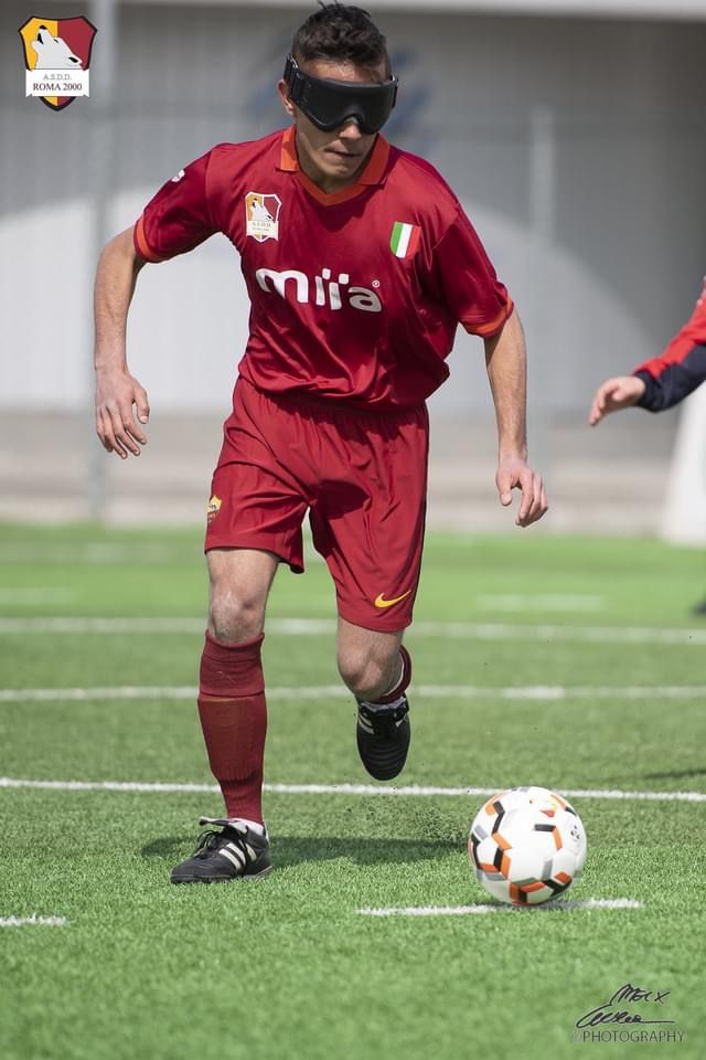 Foto torneo Città di Roma. Anno 2022. In questa foto, un calciatore della Roma Blind Football sta calciando in corsa il pallone. Indossa il completo rosso della squadra Roma Blind Football