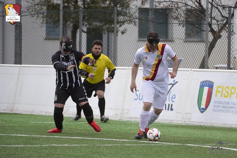 Foto Torneo di Calcio, Città di Roma. Anno 2022. In questa foto, un'atleta della Roma Blind Football sta calciando il pallone di gioco, portandolo via dal calciatore della squadra avversaria che gli sta dietro insieme all'arbitro